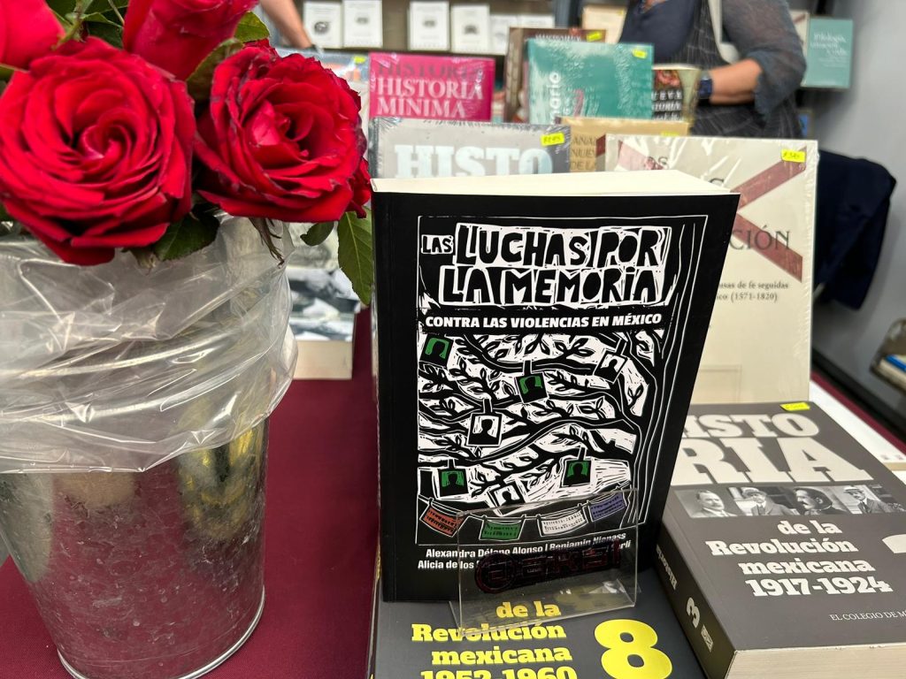 On a table with other books, next to a vase with fresh red roses, stands the book, Las Lucjas Por La Memoria (The Struggles for Memory against Violences in Mexico). The cover is an illustration of a tree with pictures of silhouetted faces clipped to its branches.
