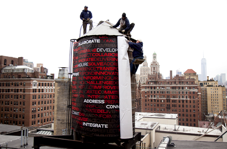 Crew members wrap the water tower on top of the building at 66 West Fifth Ave to reflect the university's new visual identity. Designed by Parsons Art Media and Technology students Rafael Cordoba and Joe Evans for their University Design Studio course. Photo by Kasia Broussalian/The New School.